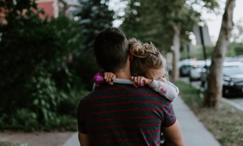 tired little girl rests her head on her father's shoulder as he carries her down the city sidewalk
